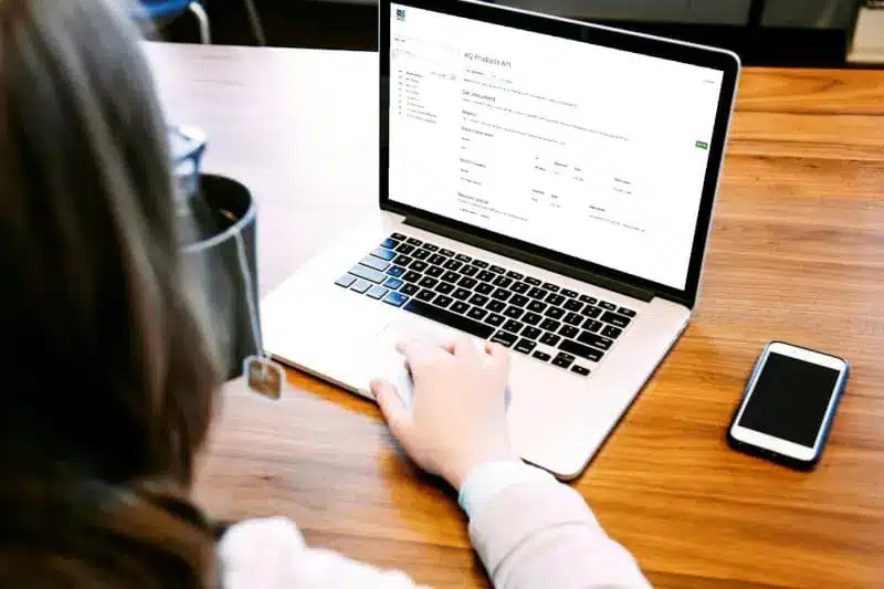 Person using a laptop at a wooden desk, viewing a website or form; a smartphone and mug with a teabag are also on the desk.