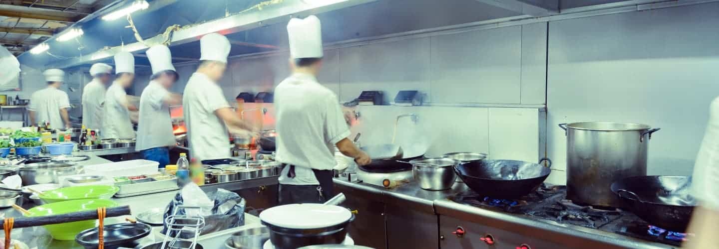 Several chefs in white uniforms and hats work at stoves in a busy commercial kitchen with various pots, pans, and cooking utensils.