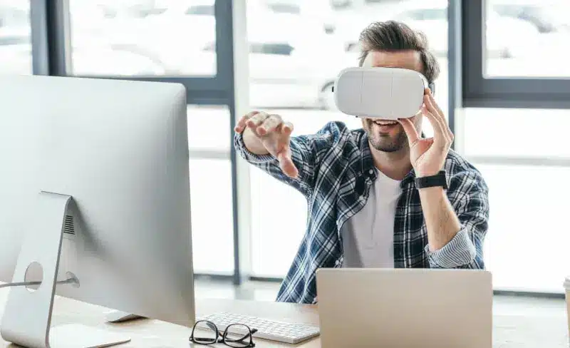 A man wearing a virtual reality headset sits at a desk with a computer, reaching forward with one hand and smiling.