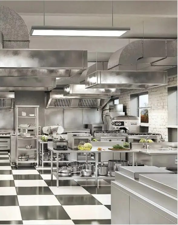 A commercial kitchen with stainless steel appliances, prep tables, and a checkered floor. Various kitchen equipment and fresh produce are visible on the counters.