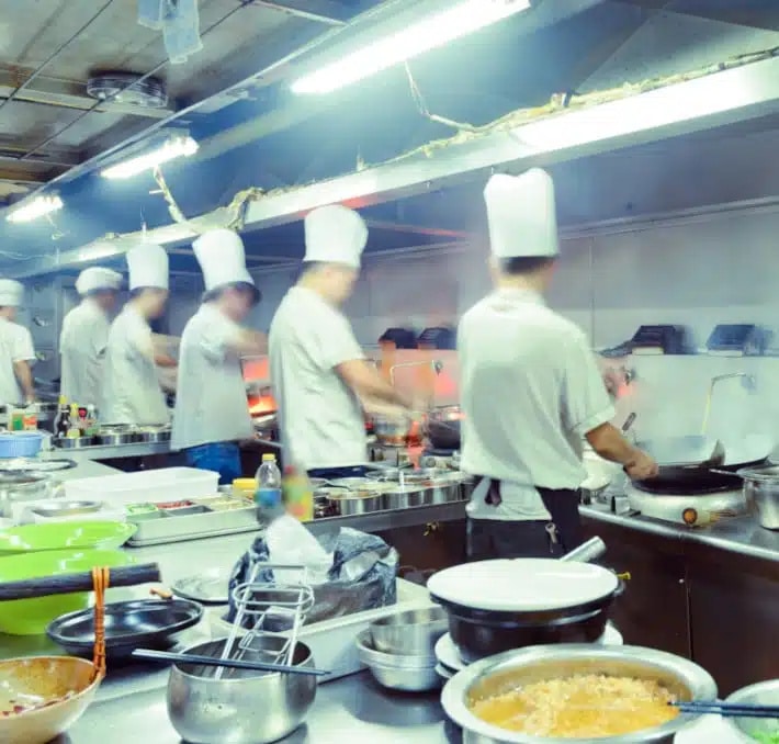 Several chefs in white uniforms and hats work in a busy, brightly lit commercial kitchen with various dishes and kitchenware visible in the foreground.