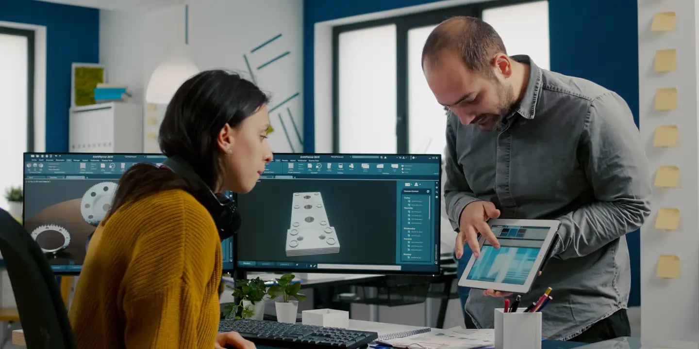 Two people in an office discuss 3D design work displayed on dual monitors and a tablet, with CAD software visible on the screens.